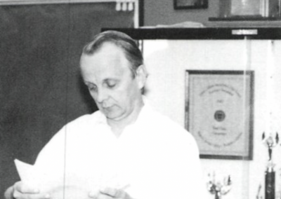 Ron Underwood reviewing papers at Beyer High School in the 1970s, standing at a podium in his classroom surrounded by student trophies and awards.